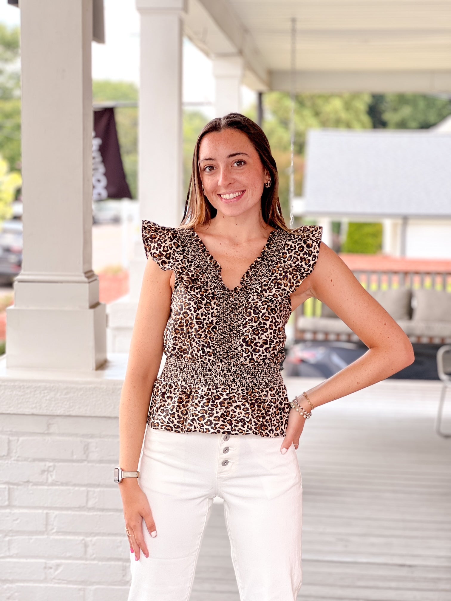 Woman wearing a leopard print top and white pants standing on a porch.